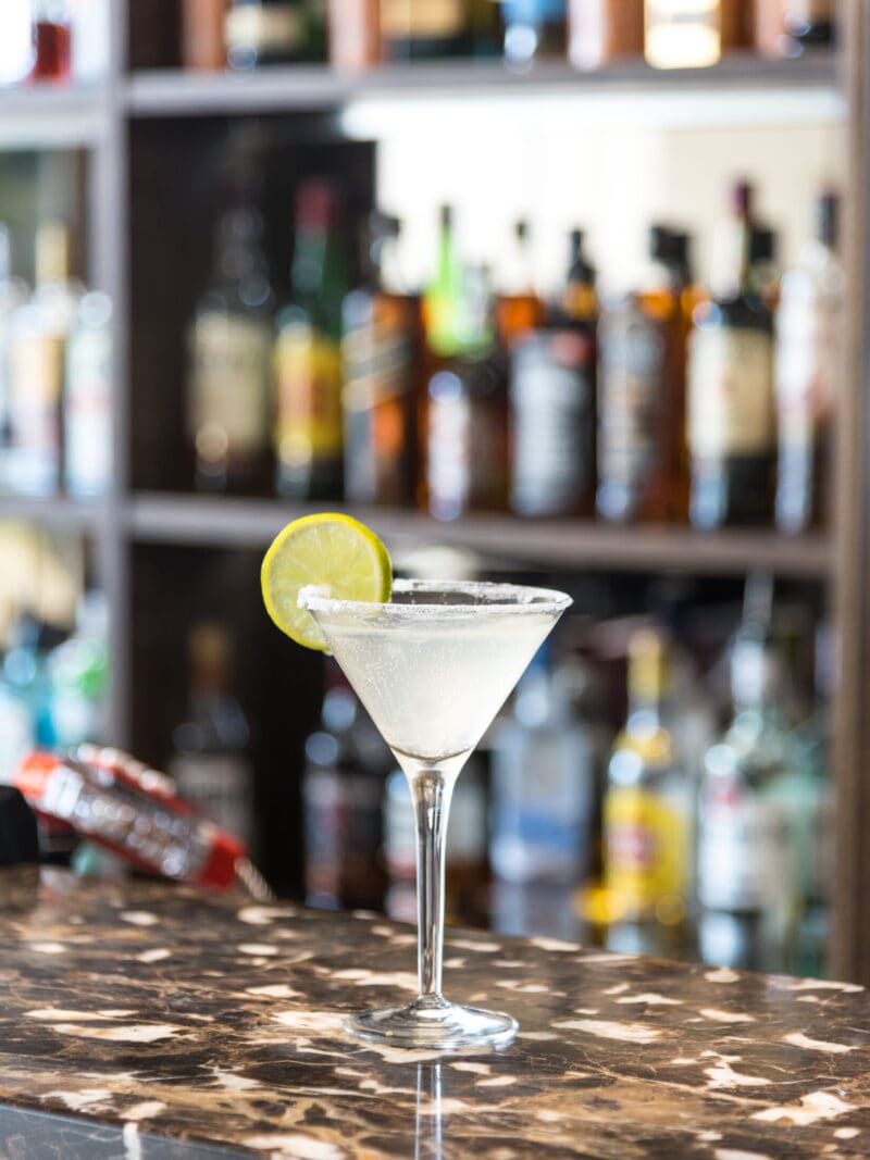 A classic Margarita or Lemon Drop cocktail served in a salted-rim martini glass on a dark marble bar counter at the Rendezvous Bar in Park Grand Heathrow. The clear, refreshing drink is garnished with a fresh lime wheel, set against a softly blurred background showcasing fully stocked liquor bottle shelves.