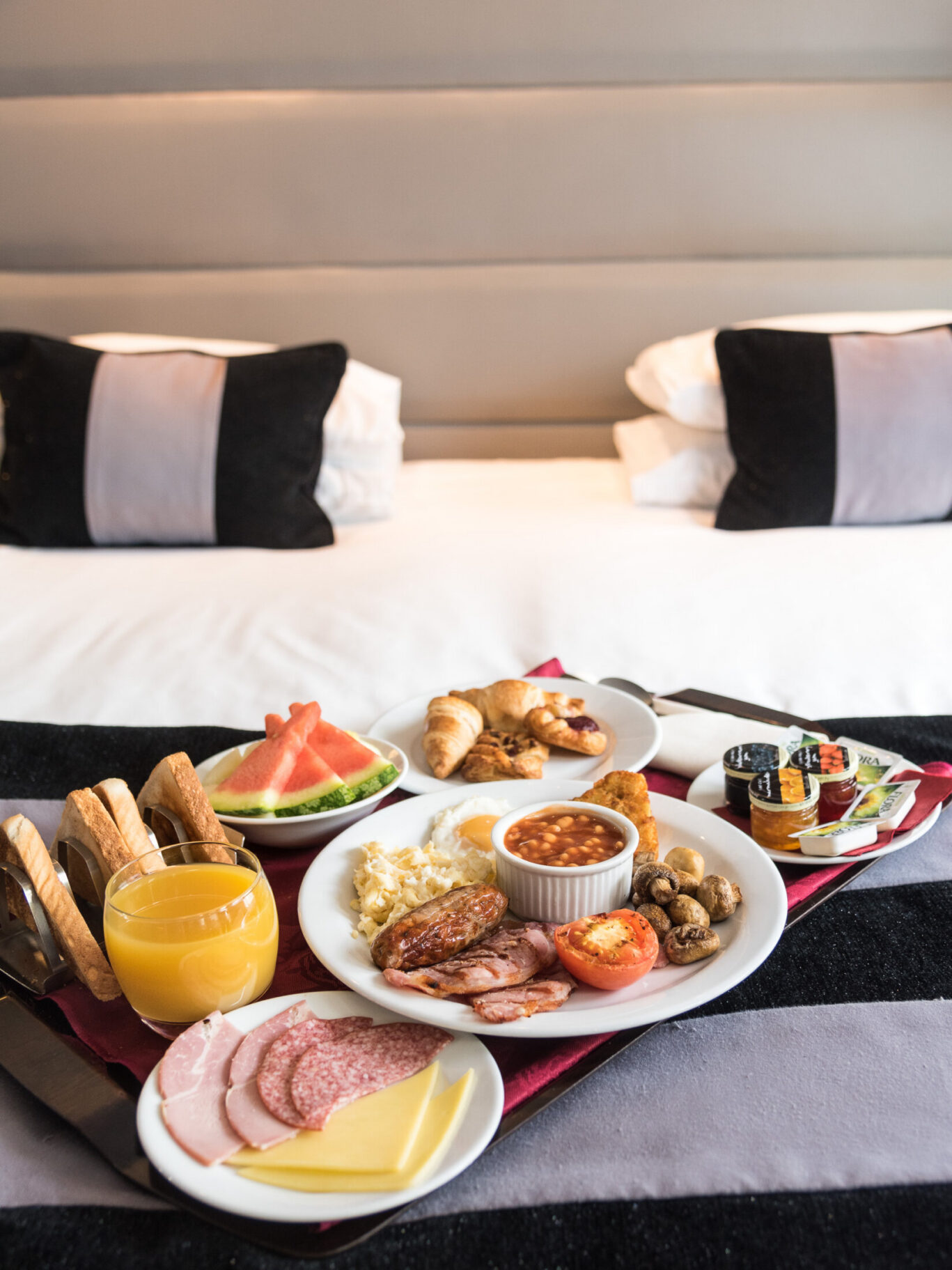 A hotel room service breakfast tray served on a bed at Park Grand Heathrow. The spread features a plate with a full English breakfast including eggs, sausage, bacon, baked beans, tomatoes, and mushrooms, accompanied by toast, fresh fruit salad, croissants, orange juice, cold cuts, and condiments.