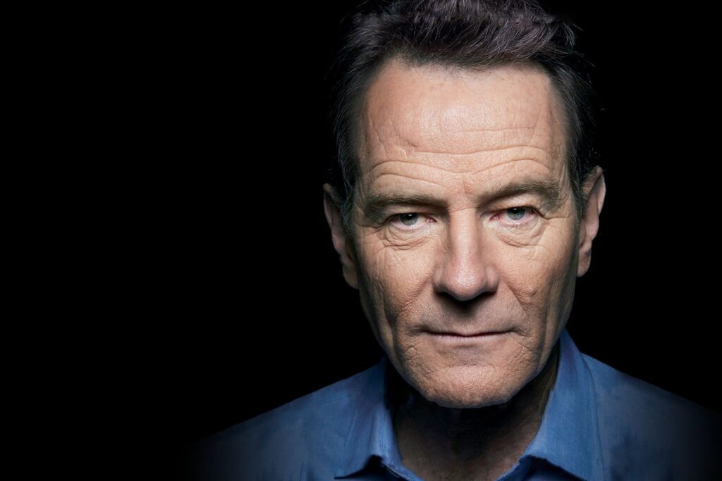 Actor Bryan Cranston stares intently at the camera in a blue unbuttoned shirt with a black background.